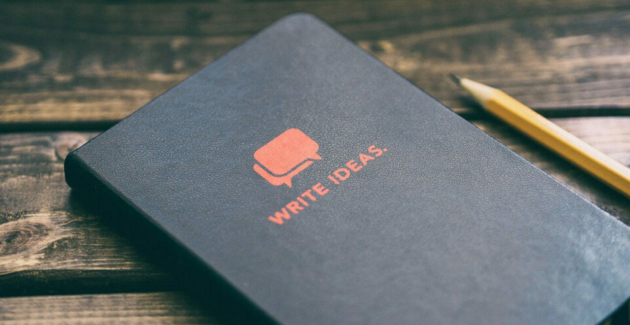 The image features a black notebook with the text "WRITE IDEAS." in orange, suggesting a space for creative thoughts or note-taking. Alongside the notebook is a yellow pencil, hinting at the readiness for writing or sketching. The items are placed on a wooden surface, which provides a warm, rustic background that complements the notebook and pencil's simple yet inviting call to creativity.