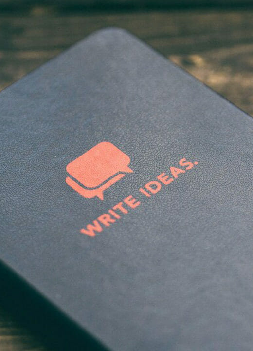 The image features a black notebook with the text "WRITE IDEAS." in orange, suggesting a space for creative thoughts or note-taking. Alongside the notebook is a yellow pencil, hinting at the readiness for writing or sketching. The items are placed on a wooden surface, which provides a warm, rustic background that complements the notebook and pencil's simple yet inviting call to creativity.